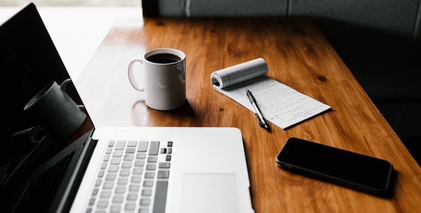 Macbook next to coffee, a notepad, and a cell phone